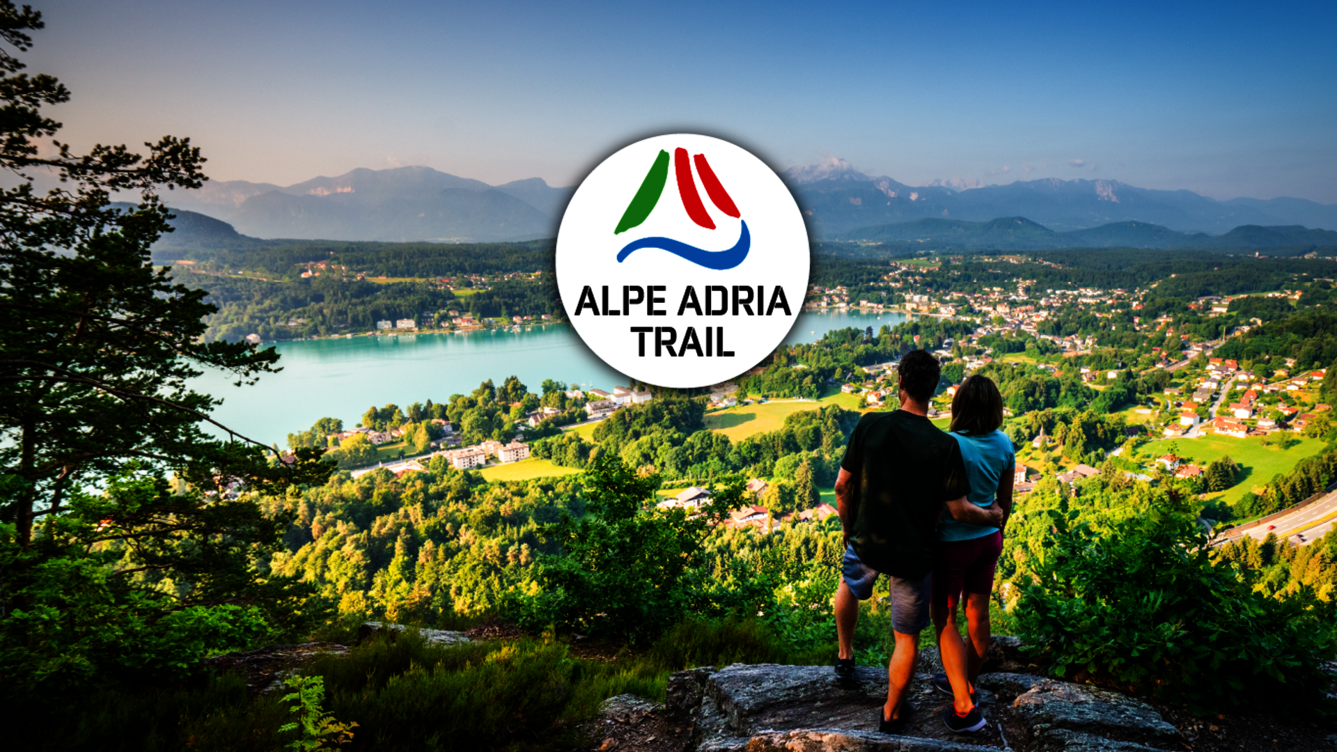 <p>Panoramaaufnahme des Wörthersee Westbeckens mit Wanderern und Logo des Alpe Adria Trail.</p>