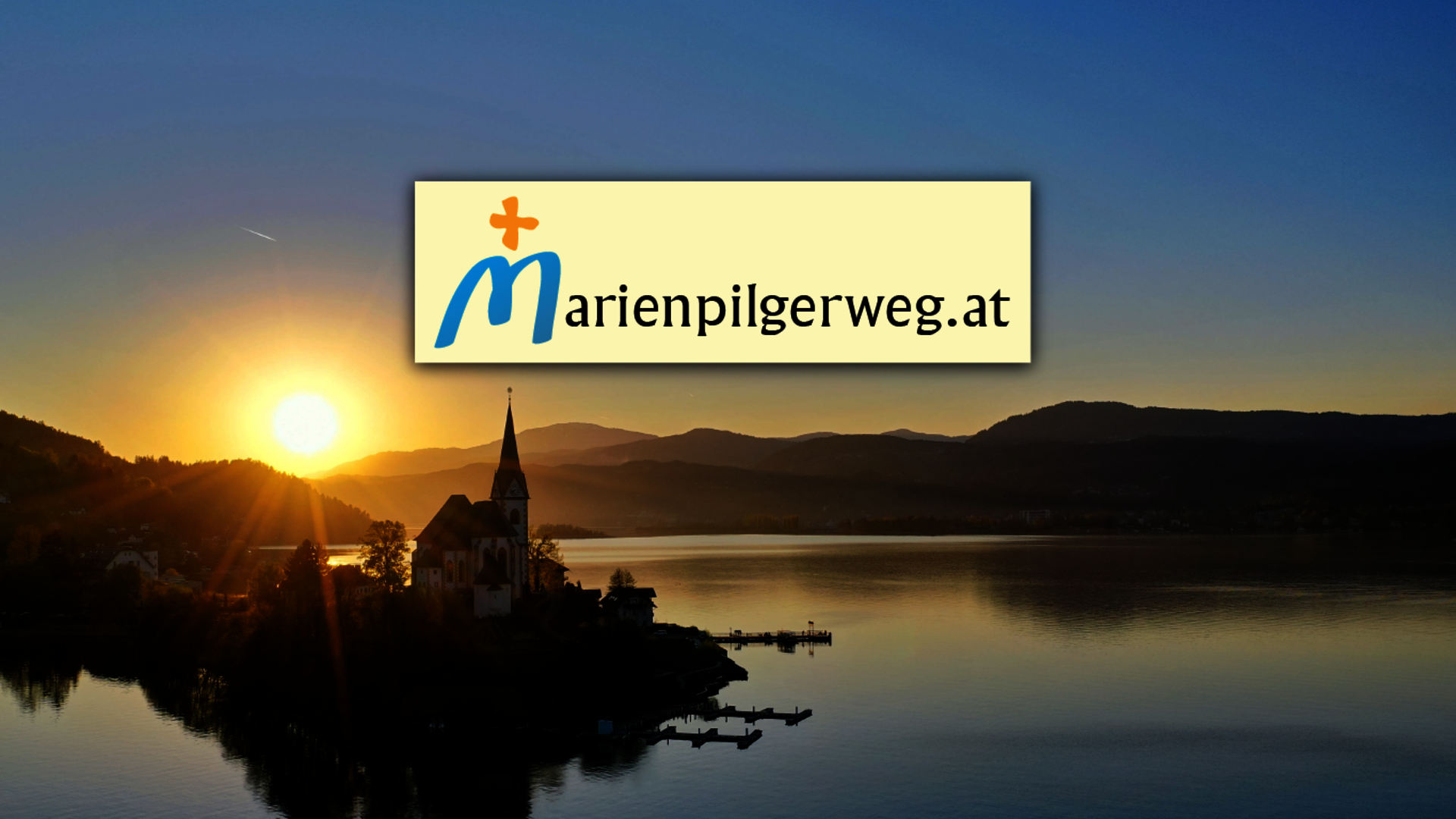 <p>Aufnahme der Kirche Maria Wörth im Sonnenuntergang mit Logo des Marienpilgerweges.</p>