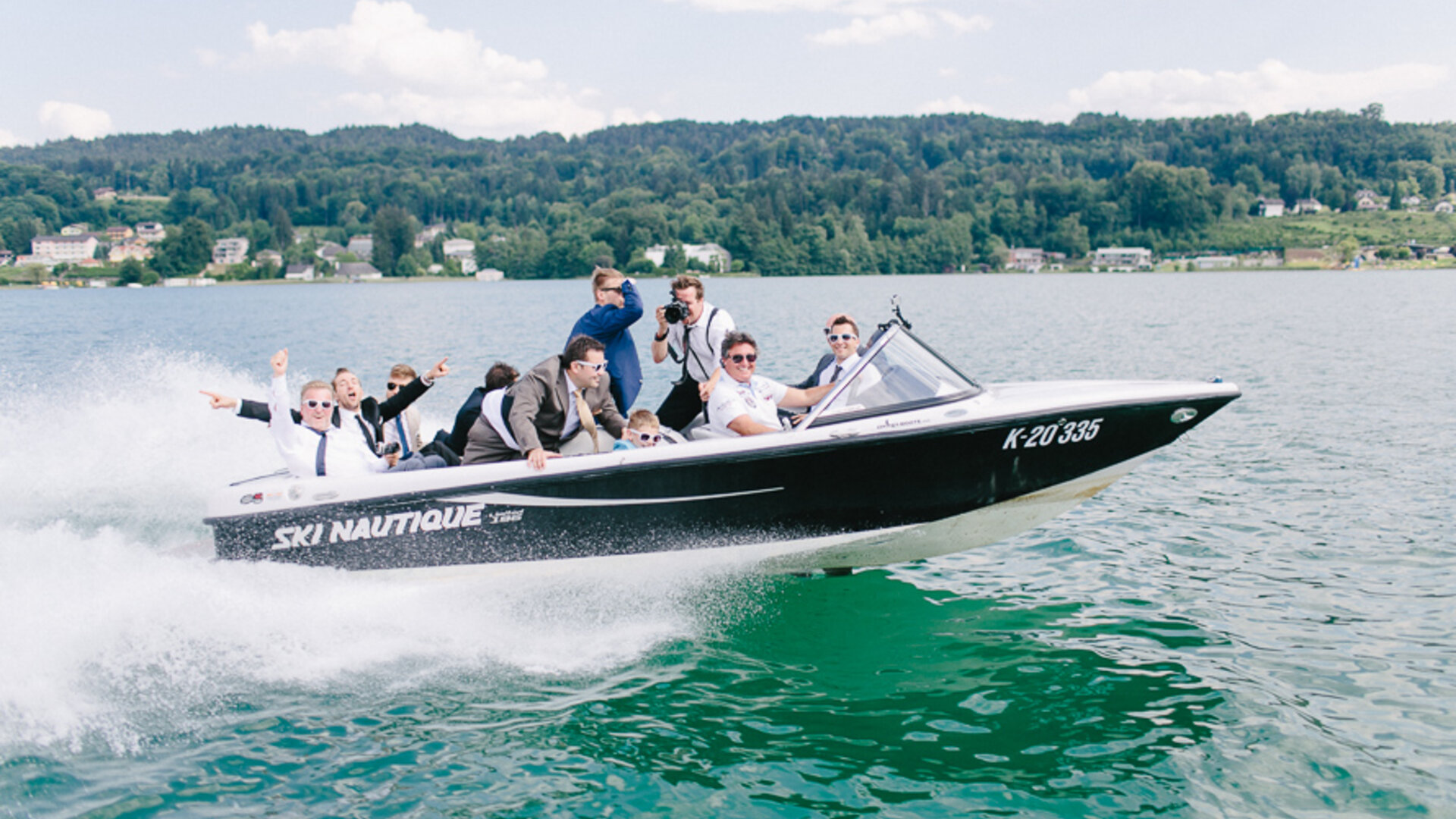 <p>Gruppe von jungen Männern als Partygesellschaft auf einem gleitenden Motorboot im türkisen Wörthersee.</p>